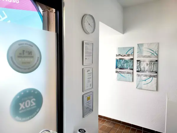 Heller Eingangsbereich mit Auszeichnungen an der Tür, moderner Uhr und abstrakter Wandkunst.